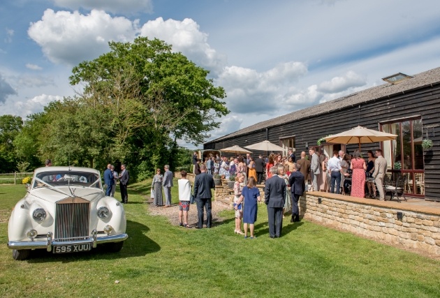 outdoors terrace adjacent to barn, people milling about wedding car on grass sunny day