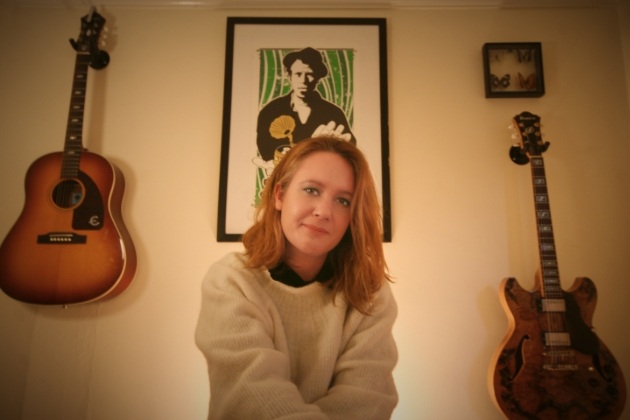 woman sitting down with two guitars hanging up behind her