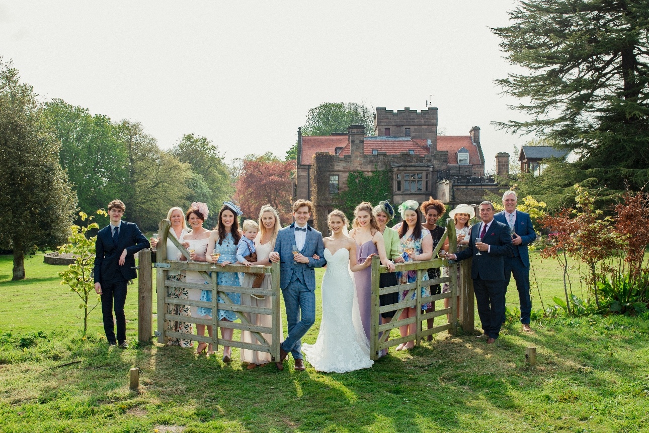 bride and groom and their guest posing at a field gate with manor house in the background