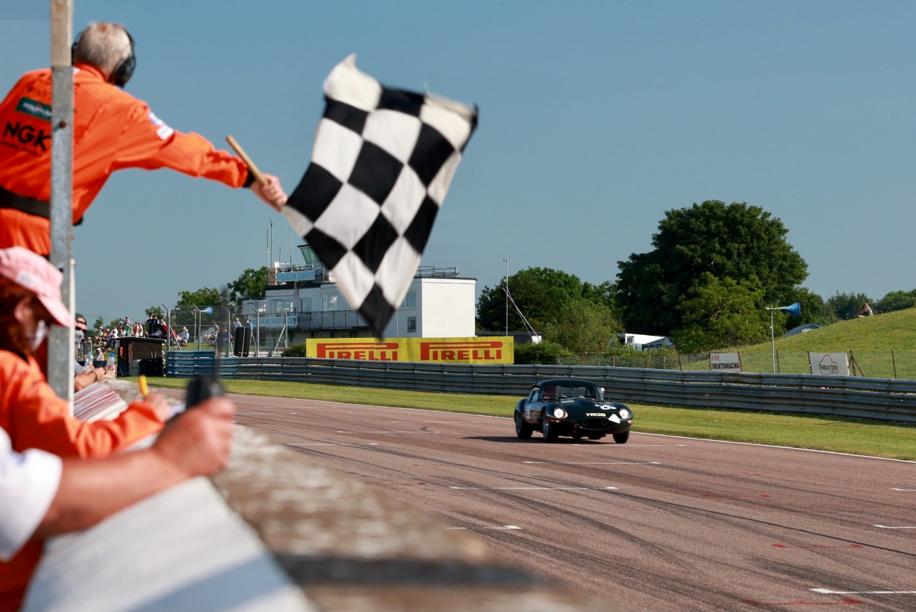 black retro race car, on a track with a linesman with a finish flag and stands in the backfground