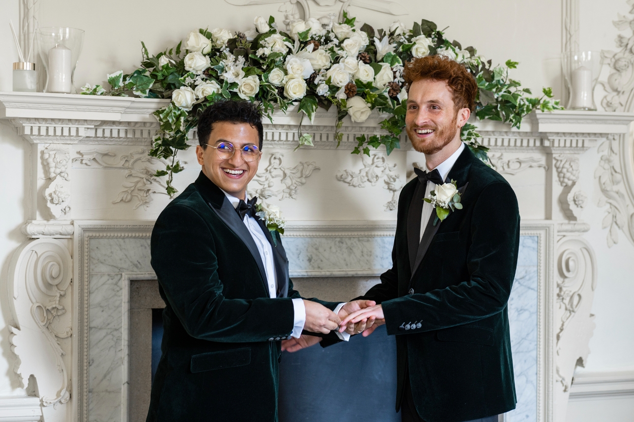two grooms in black velvet suits wed in front of a stately home white fireplace