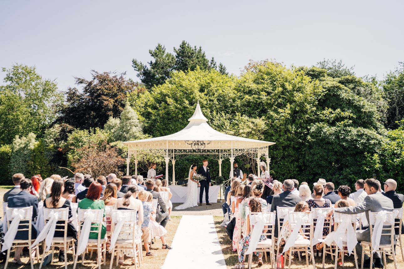 white wedding pavillion in garden couple tying the knot under it, guests outside