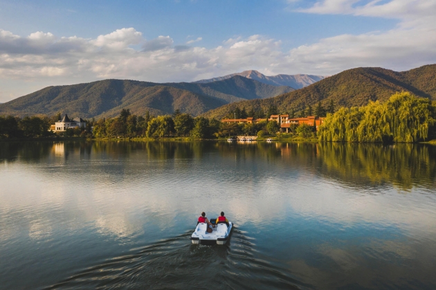 A large lake with mountains in the distance with two people on a boat heading towards a building on the shore