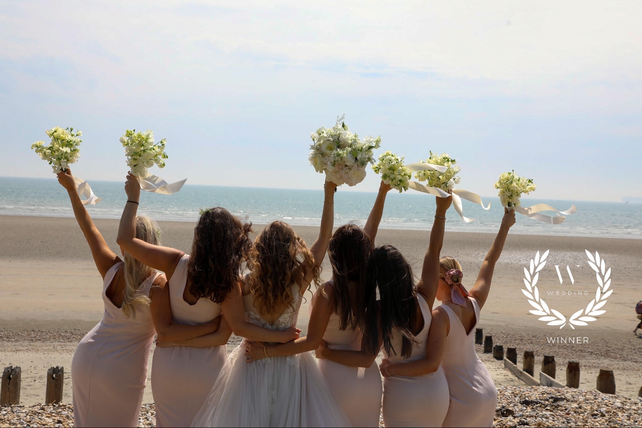 bride and bridesmaide on a beach holding flowers in hair back to camera looking out to sea